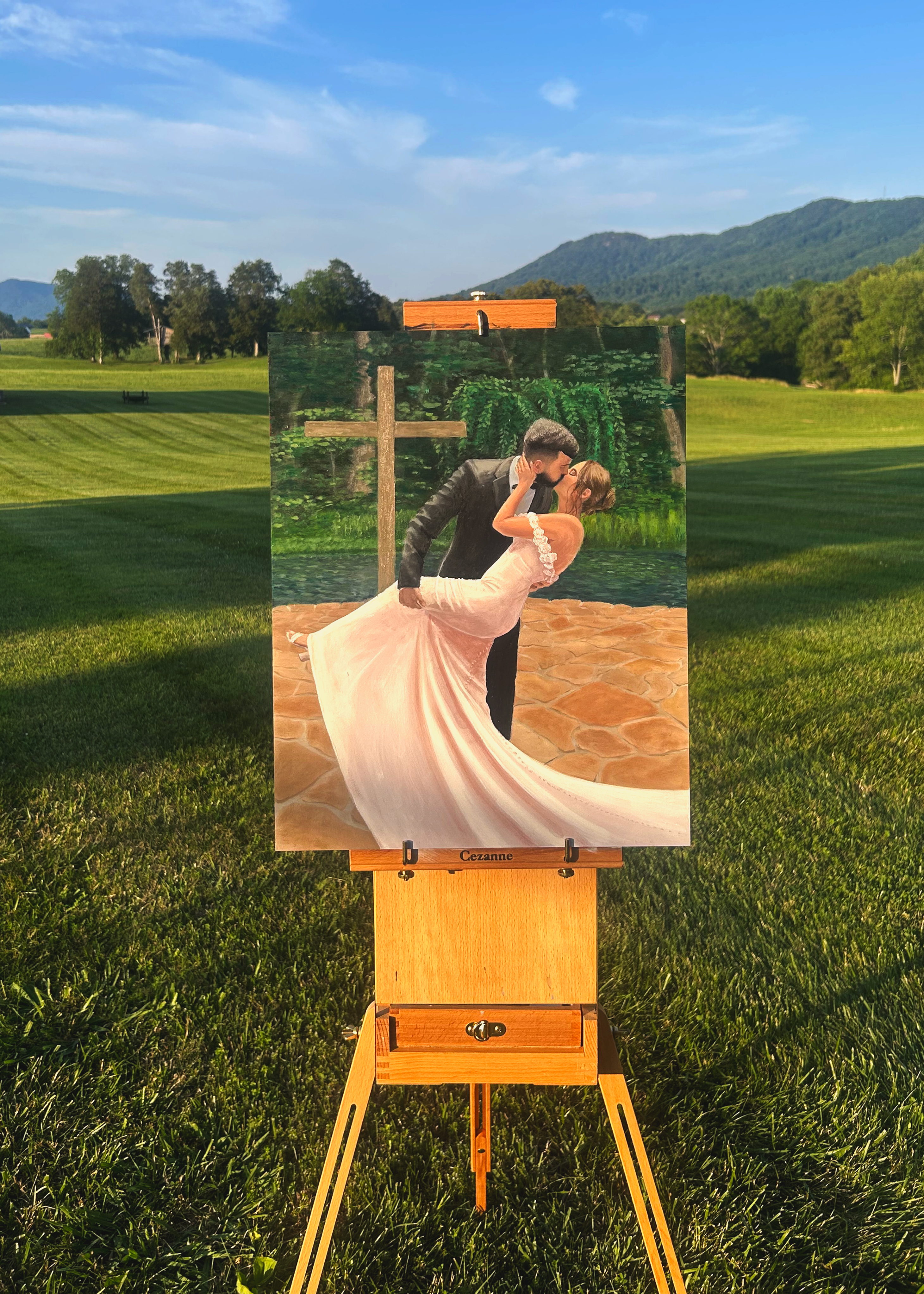 Live wedding painting in oil of a bride and groom's dip kiss, on an easel outdoors with mountains behind